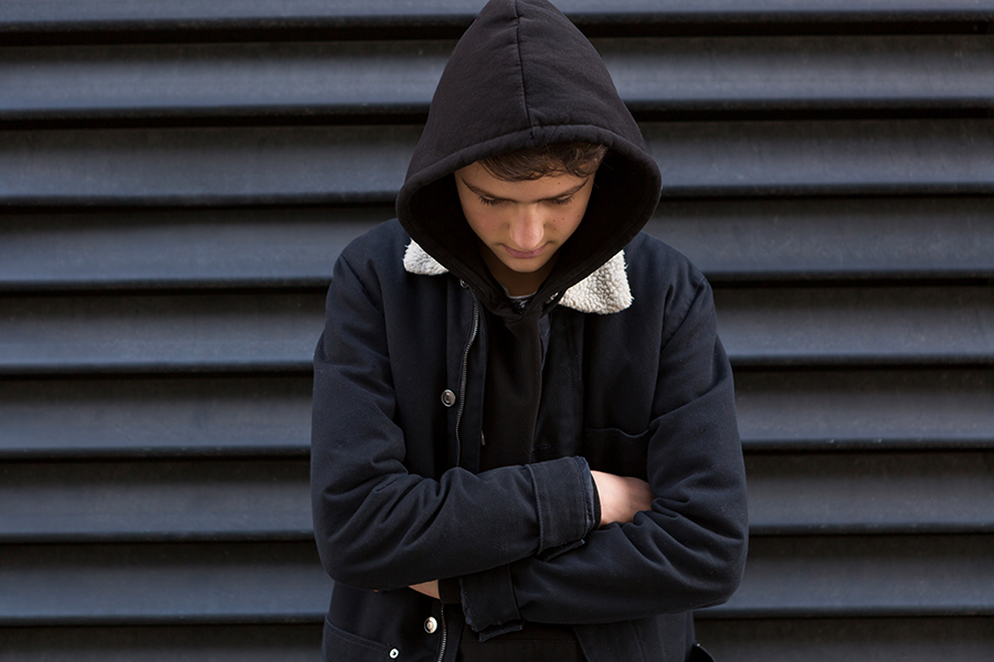 Ein Jugendlicher mit einem schwarzen Kapuzenpullover und gesenktem Kopf steht vor geschlossenen Jalousien.