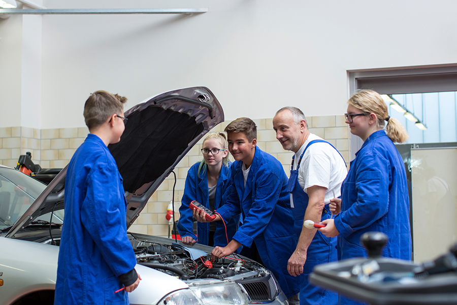 Ein Ausbilder sowie 2 Schüler und 2 Schülerinnen stehen an einem Auto mit offener Motorhaube.